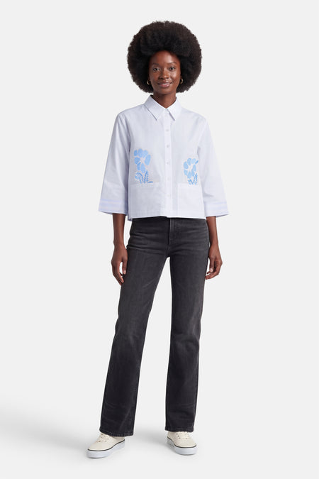 Chemise blanche de MORE & MORE à manches trois-quarts avec broderies florales bleues sur les poches et poignets rayés, portée avec un jean.