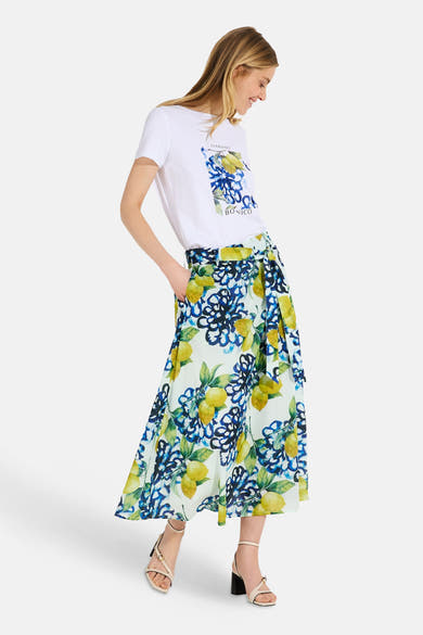 Jupe longue multicolore de MORE & MORE, imprimé floral et citrons, portée avec un t-shirt blanc et des sandales beiges à talons.
