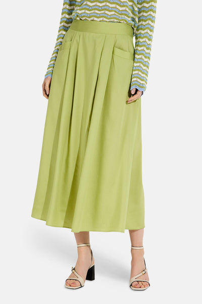 Jupe longue verte de MORE & MORE, plissée avec des poches, portée avec un pull à motifs et des sandales à talons.
