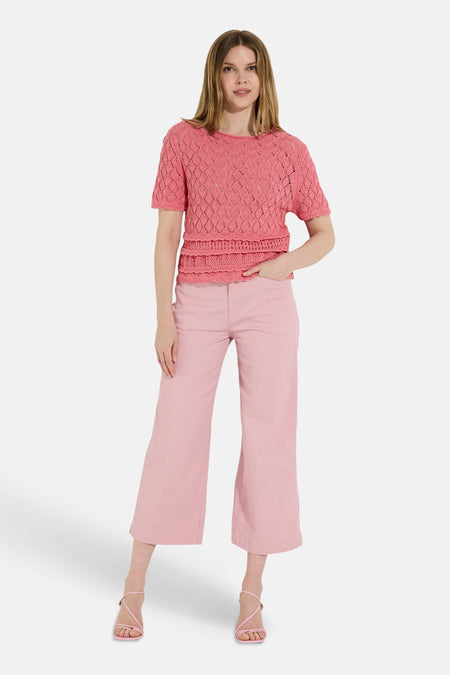 Pull rose à manches courtes de MORE & MORE, tricoté avec un motif ajouré et ourlet à volants, porté avec un pantalon rose.
