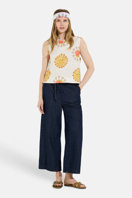 Blouse sans manches multicolore de MORE & MORE, à motifs de soleils jaunes et fleurs roses, portée avec un jean large et un bandeau assorti.