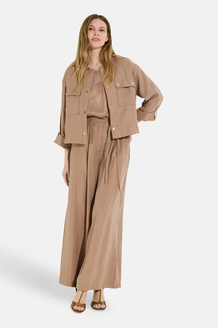 Pantalon de costume brun de MORE & MORE, à coupe large et fluide, porté avec un chemisier et une veste assortis, et des sandales.