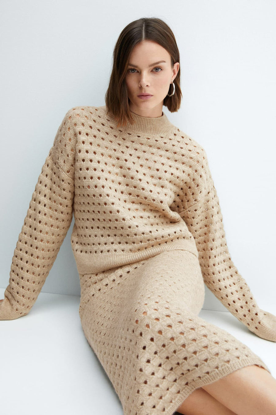 Pull beige de MANGO, à motif ajouré, porté avec une jupe assortie et des créoles.