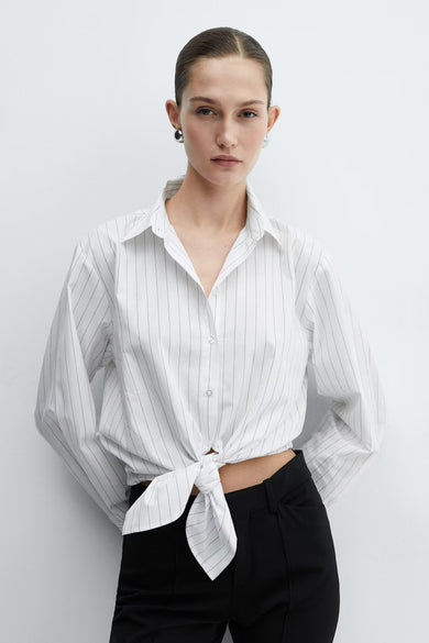 Chemise à manches longues blanche à rayures by MANGO, nouée à la taille, portée avec un pantalon noir.