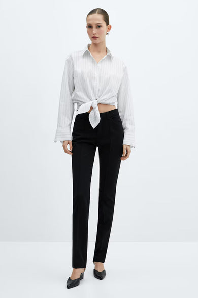 Chemise à manches longues blanche rayée par MANGO, nouée à la taille, portée avec un pantalon noir et des mules.
