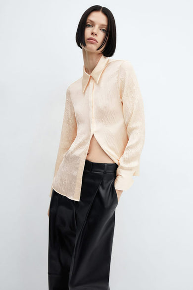 Chemise à manches longues beige de MANGO, avec une texture froissée et portée ouverte sur un pantalon noir.
