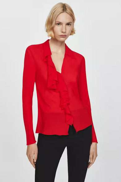 Blouse rouge à manches longues par MANGO, avec des volants sur le devant et portée avec un pantalon noir.

