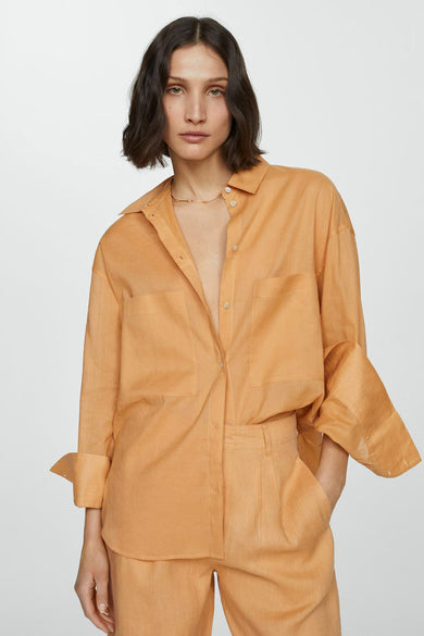 Chemise à manches longues orange de MANGO, portée ouverte avec un pantalon assorti et un collier doré.