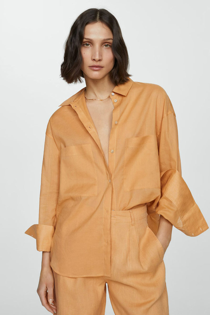 Chemise à manches longues orange de MANGO, portée ouverte avec un pantalon assorti et un collier doré.