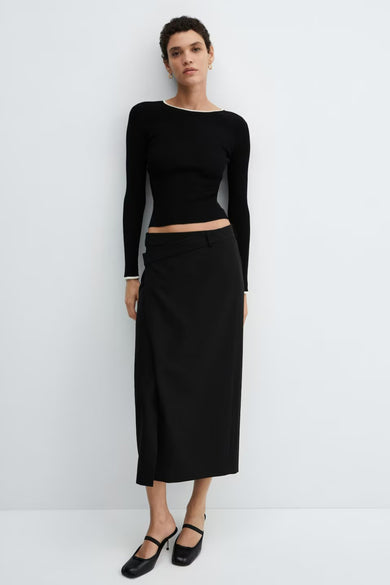 Pull noir à manches longues de MANGO, porté avec une jupe noire fendue et des mules à talons.