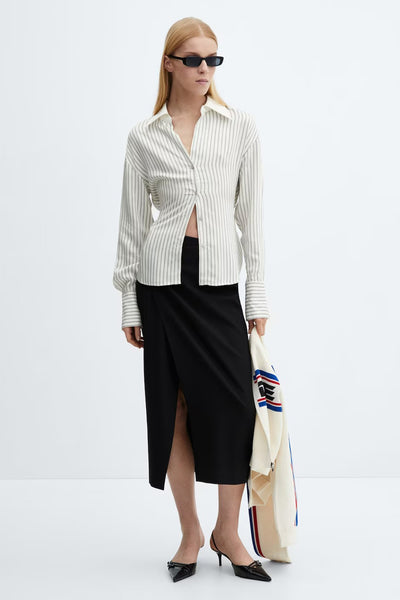 Chemisier beige à rayures verticales de MANGO, porté avec une jupe noire fendue et des lunettes de soleil.