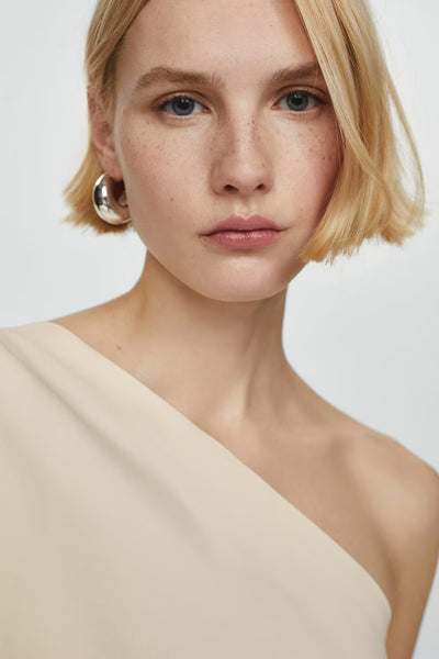 Blouse beige de MANGO, coupe asymétrique, portée avec une boucle d'oreille argentée.