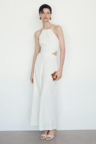 Combinaison beige de MANGO, avec découpe à la taille, tenue avec une pochette dorée et des chaussures blanches.