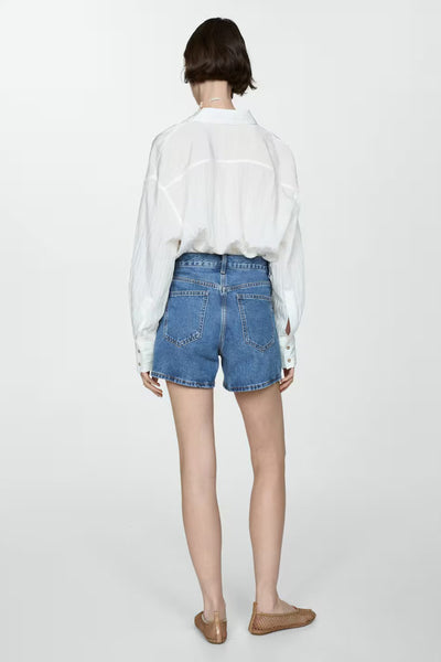 Short en denim bleu par MANGO, porté avec une chemise blanche ample et des chaussures plates marron.