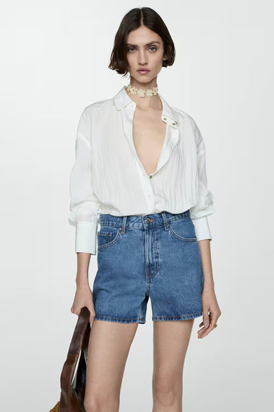 Short en denim bleu clair de MANGO, porté avec une chemise blanche ouverte et un collier à fleurs.
