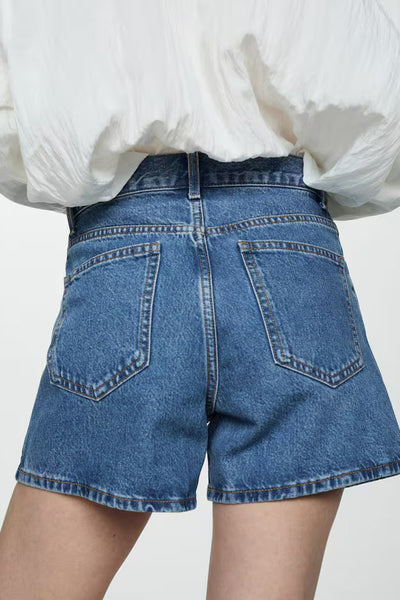 Short en denim bleu de MANGO, avec poches arrière et ourlet effiloché, porté avec un chemisier blanc.