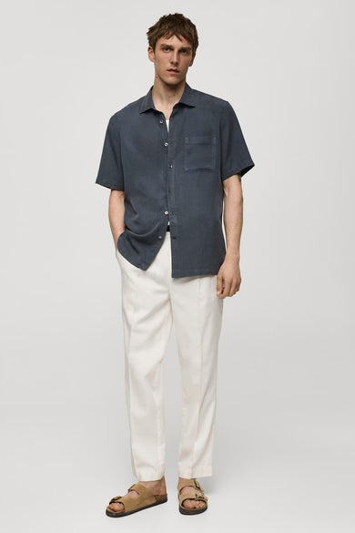 Chemise à manches courtes bleue de MANGO, portée avec un pantalon blanc et des sandales marron clair.