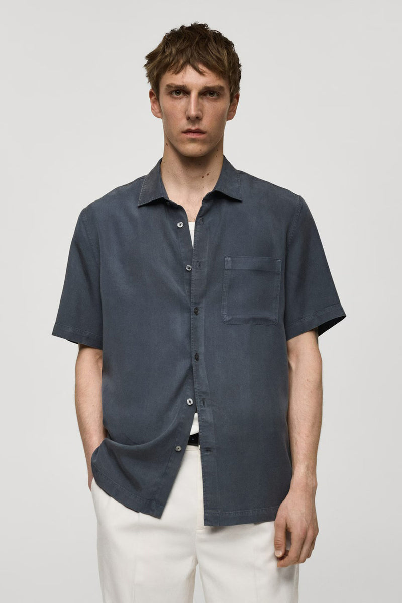 Chemise bleue à manches courtes de MANGO, avec poche poitrine, portée avec un pantalon blanc.