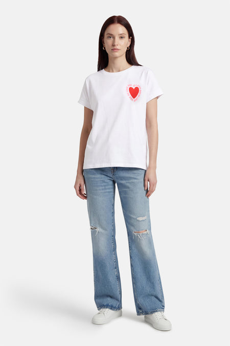 T-shirt blanc de Terra di Siena, orné d'un cœur rouge et d'un texte rose, porté avec un jean et des baskets.
