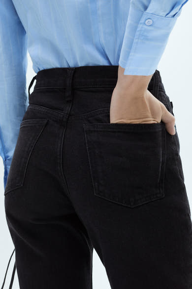 Zwarte 'Mom' jeans van MANGO, met een hand in de achterzak, gedragen met een blauw gestreept overhemd.