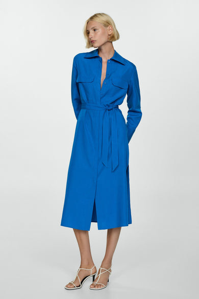 Blauwe jurk met lange mouwen van MANGO, met een strikceintuur en witte sandalen met hak.