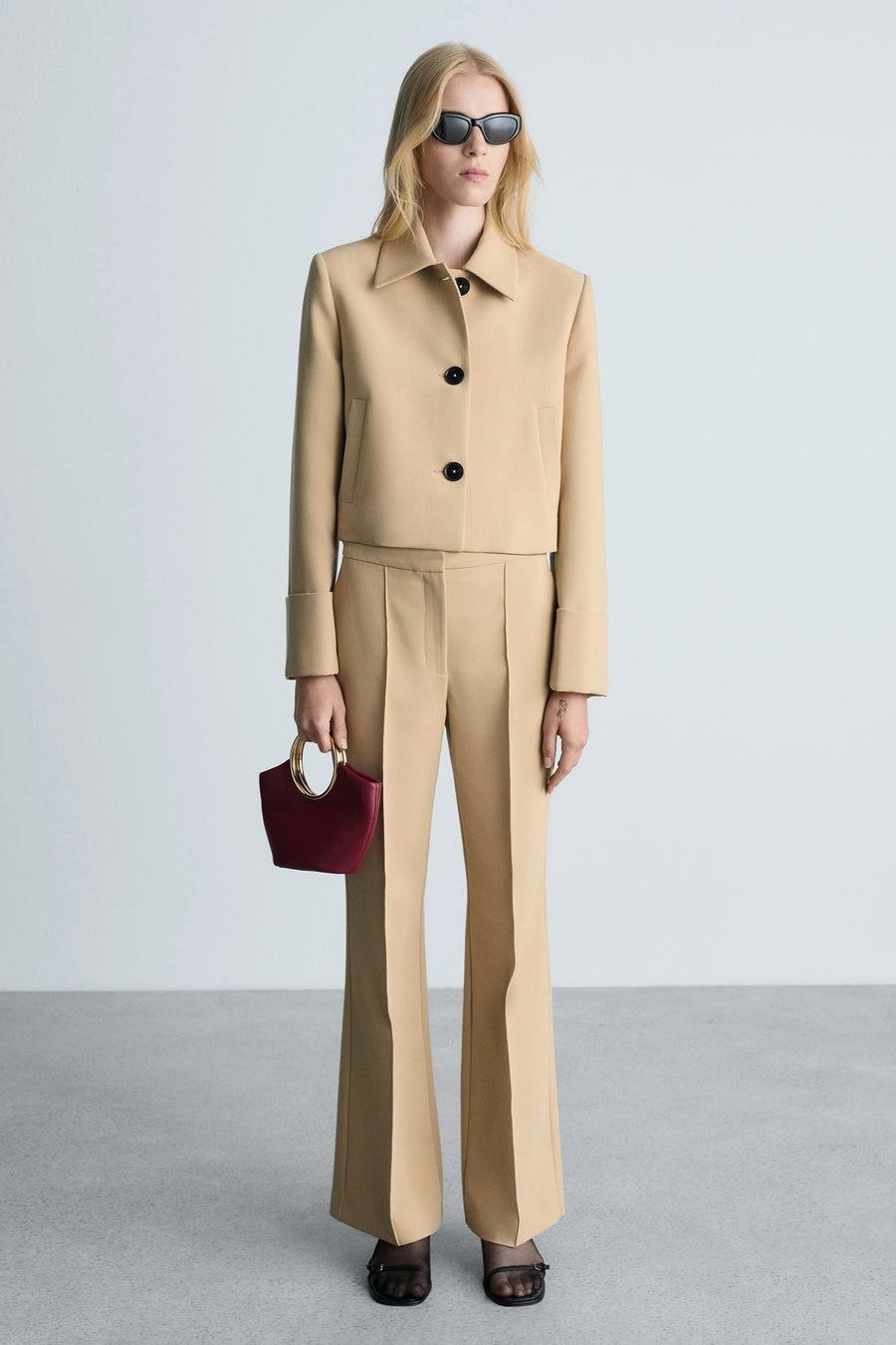 Bruine broek van MANGO, uitlopende pasvorm, gedragen met een bijpassend beige jasje en een bordeauxrode tas met gouden hengsel.
