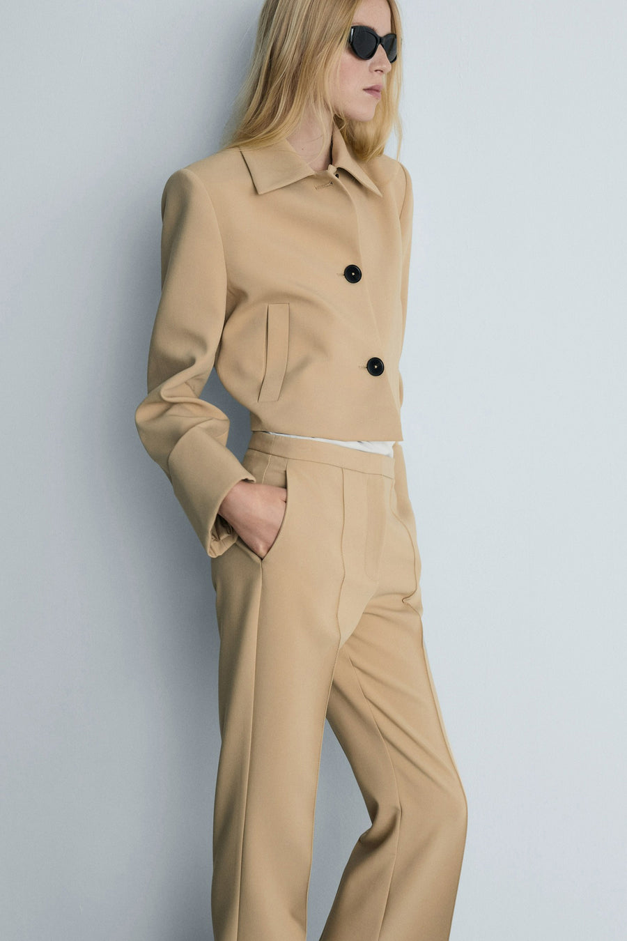 Bruine broek van MANGO, rechte pasvorm, gedragen met een korte beige jas en een zwarte zonnebril.