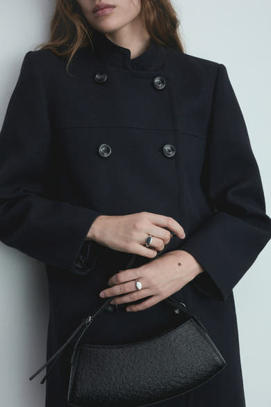 Veste longue bleue de MANGO, avec boutons, portée avec un sac à main texturé et des bagues argentées.
