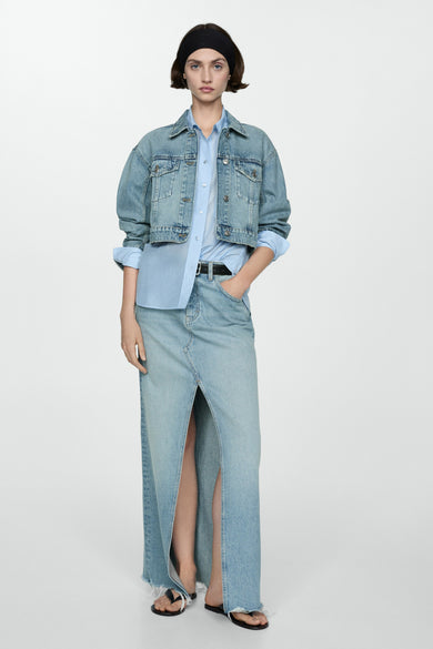Veste en denim bleu moyen de MANGO, courte avec un chemisier bleu clair, jupe maxi en denim fendu et sandales.