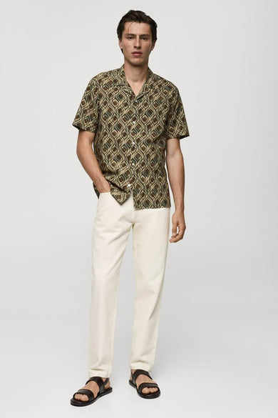Chemise à manches courtes verte à motifs, de MANGO, portée avec un pantalon blanc et des sandales noires.