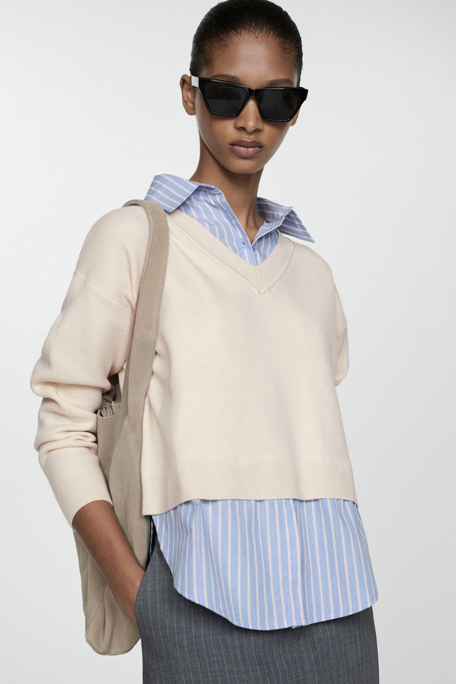 Beige V-hals trui van MANGO, gedragen over een gestreept blauw overhemd en een gestreepte grijze broek, met een tas.