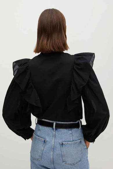 Chemisier noir à manches longues par MANGO, avec des volants aux épaules, porté avec un jean bleu et une ceinture noire.