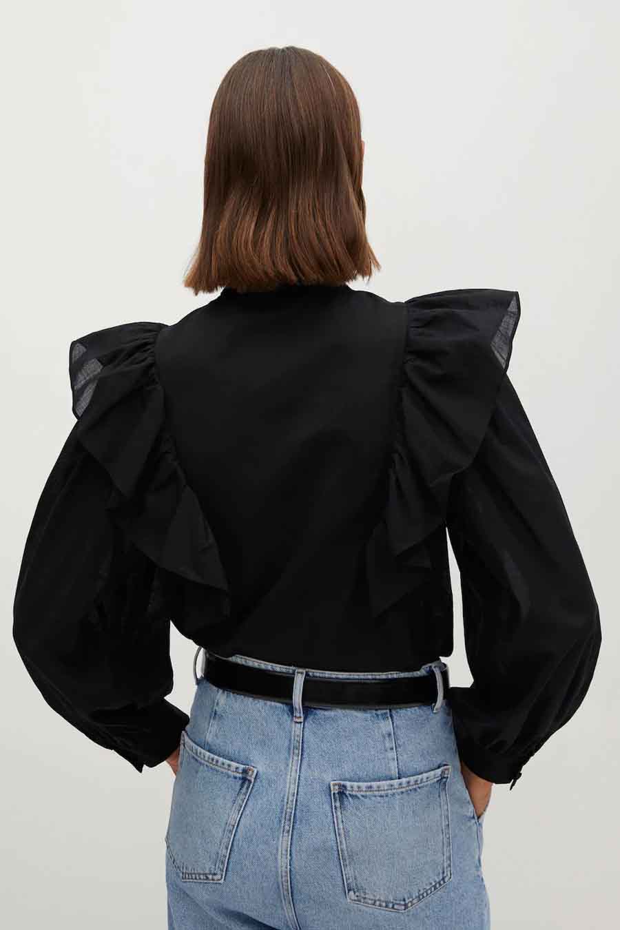 Chemisier noir à manches longues par MANGO, avec des volants aux épaules, porté avec un jean bleu et une ceinture noire.