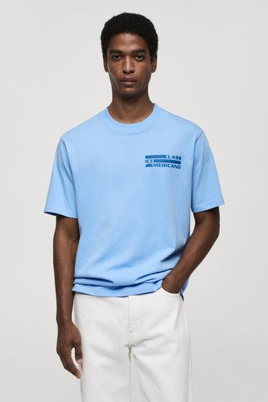 T-shirt bleu à manches courtes de MANGO, avec imprimé graphique sur le devant, porté avec un pantalon blanc.
