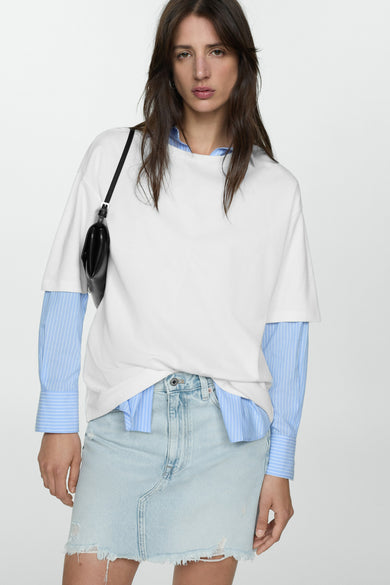 Jupe courte bleu denim by MANGO, ourlet effiloché, portée avec un t-shirt blanc et une chemise rayée bleue.