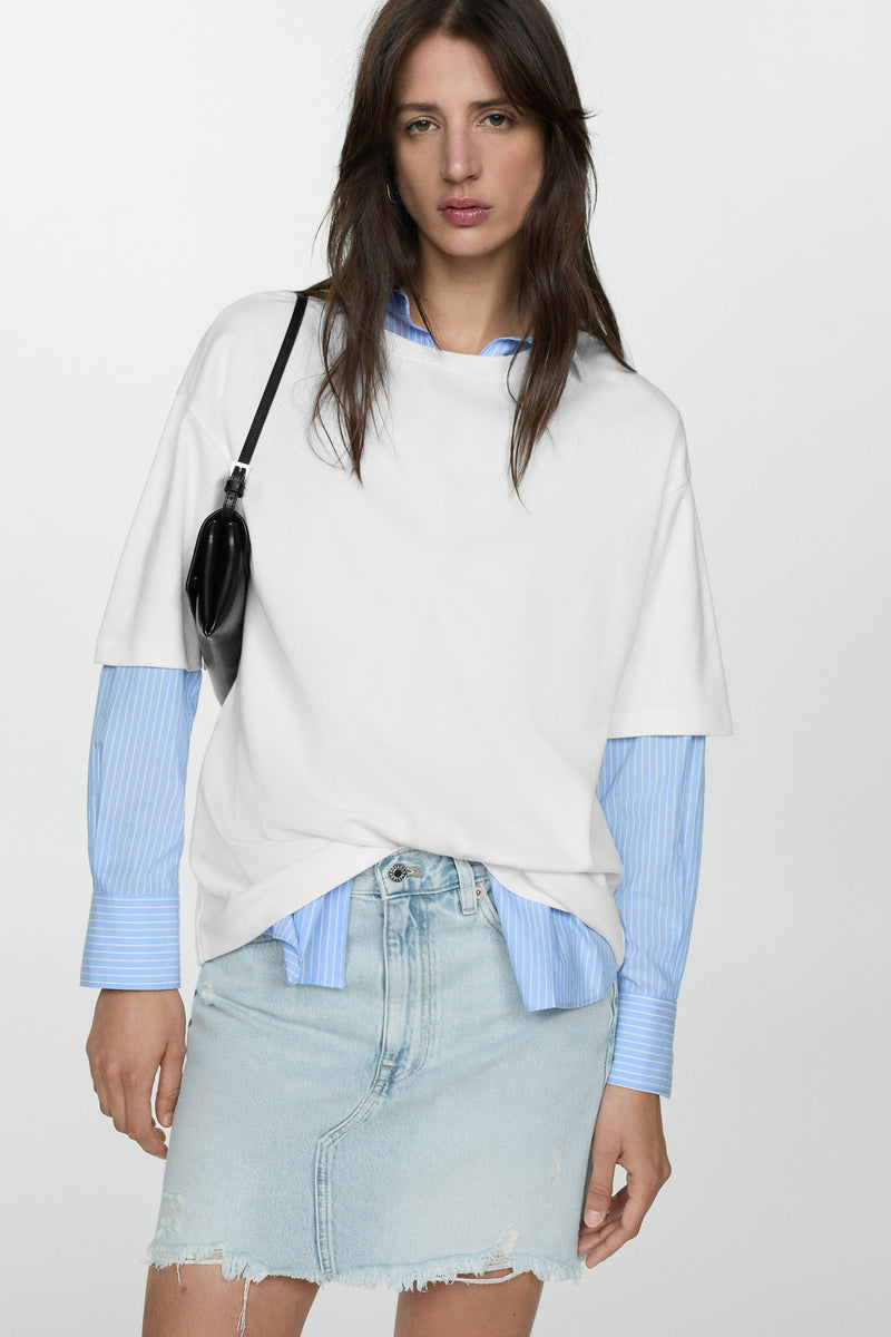 Jupe courte bleu denim by MANGO, ourlet effiloché, portée avec un t-shirt blanc et une chemise rayée bleue.
