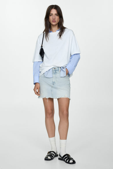 Jupe courte en denim bleu moyen de MANGO, avec ourlet effiloché, portée avec une chemise rayée et des sandales.