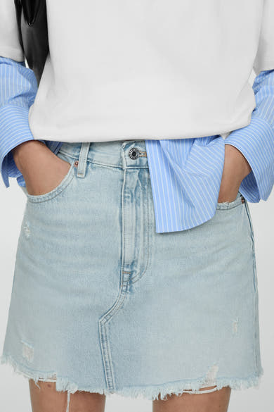 Korte spijkerrok in middieblauw van MANGO, met rafelige randen en opgestikte zakken, gedragen met een wit T-shirt en een blauw gestreept overhemd.