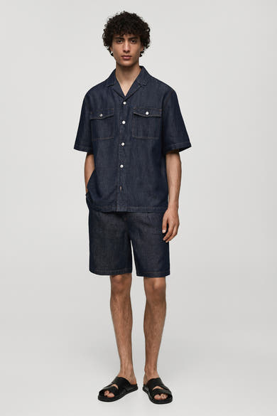 Short en denim bleu moyen de MANGO, porté avec une chemise assortie et des sandales en cuir noir.