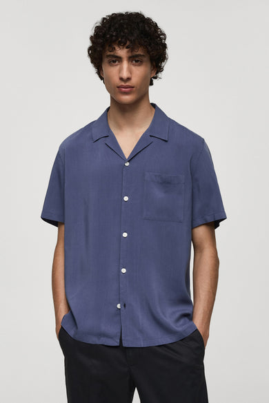 Chemise bleue à manches courtes par MANGO, avec poche de poitrine et boutons blancs, portée avec un pantalon noir.

