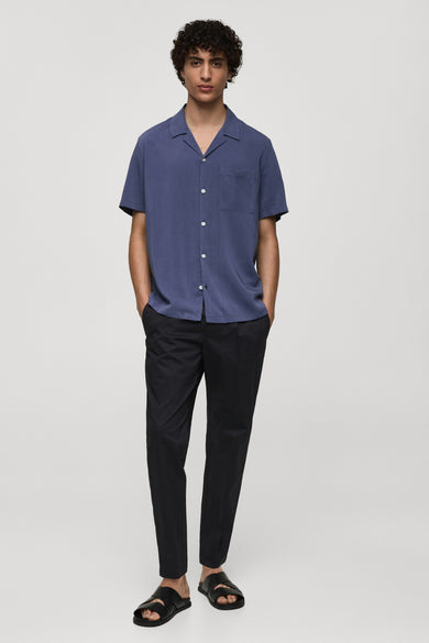 Chemise bleue à manches courtes de MANGO, avec poche poitrine et boutons blancs, portée avec un pantalon noir et des sandales.
