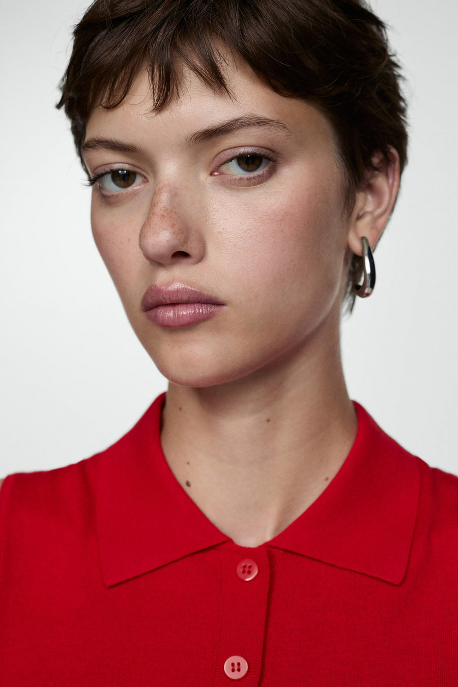 Rode polo van MANGO, geknoopt, gedragen met zilveren oorbellen en discrete make-up.