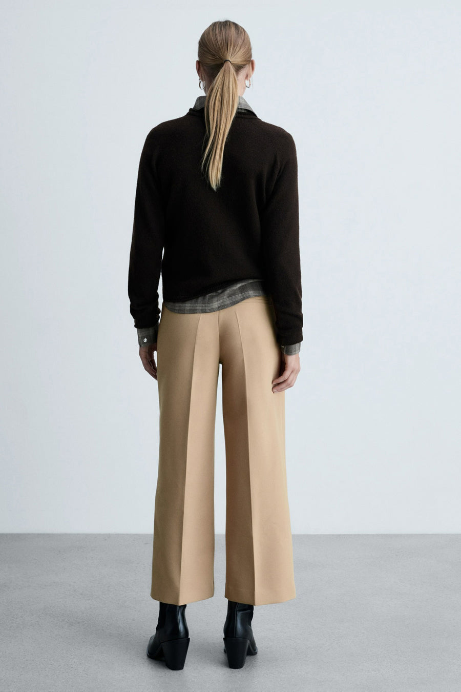 Beige broek van MANGO, gedragen met een bruine trui en zwarte enkellaarsjes, van achteren gezien.