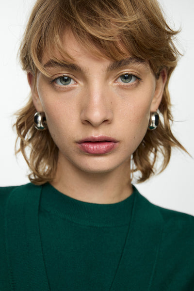 Gilet vert de MANGO, porté avec un pull vert foncé et des boucles d'oreilles argentées.