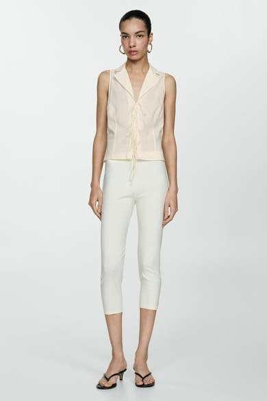 Blouse beige sans manches de MANGO, à col tailleur et fermeture à lacets, portée avec un pantalon capri blanc.
