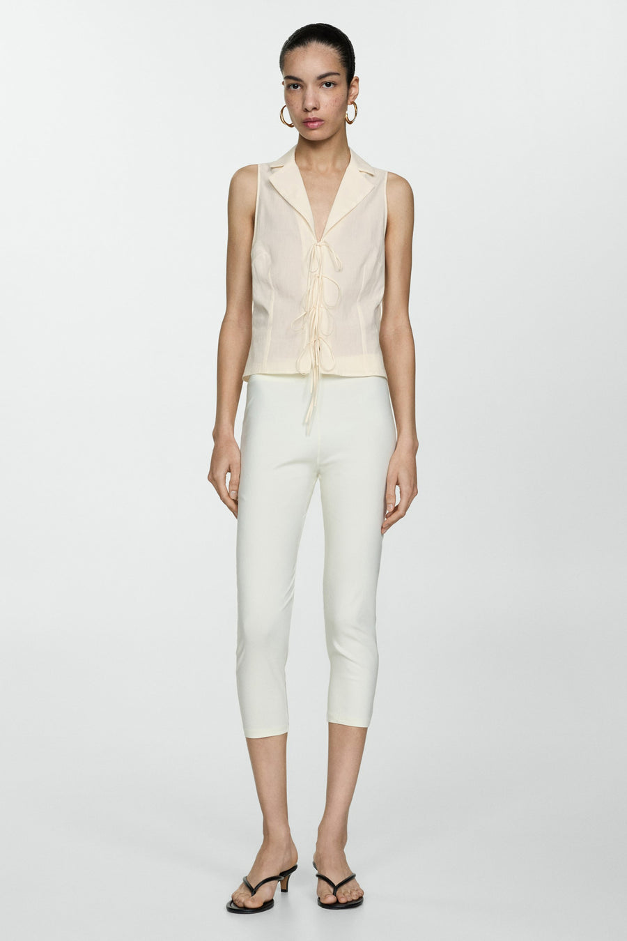Blouse beige sans manches de MANGO, à col tailleur et fermeture à lacets, portée avec un pantalon capri blanc.
