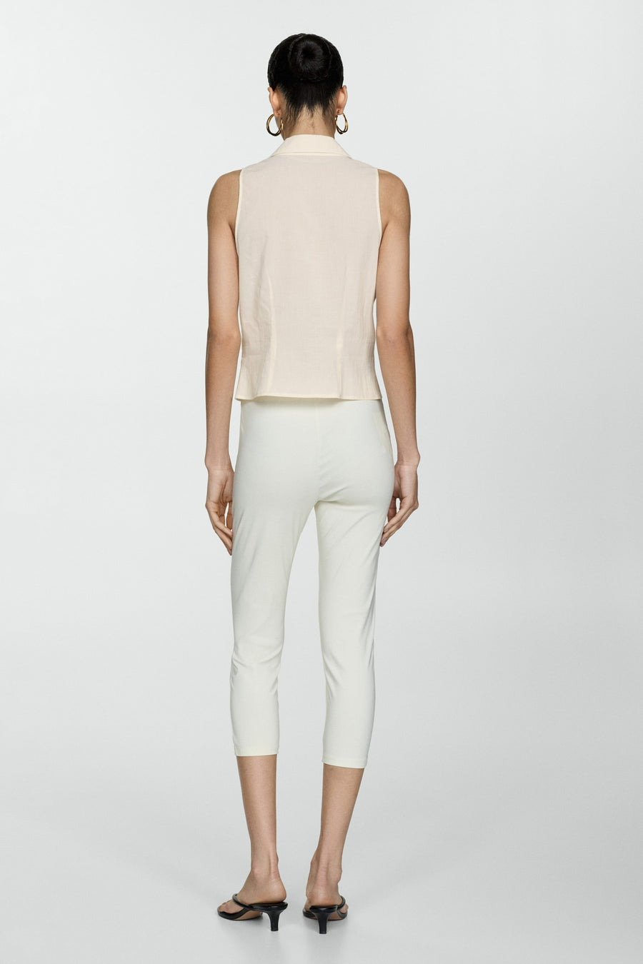 Chemisier beige sans manches de MANGO, porté avec un pantalon blanc court et des sandales noires à talons aiguilles.