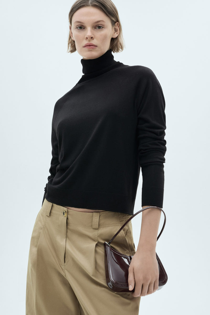 Zwarte rolluikensjas van MANGO, gedragen met een beige broek en een klein rode handtas.