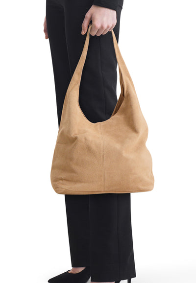 Ecru suede shoulder bag with soft slouchy design, carried by model in black pants and shoes, side view