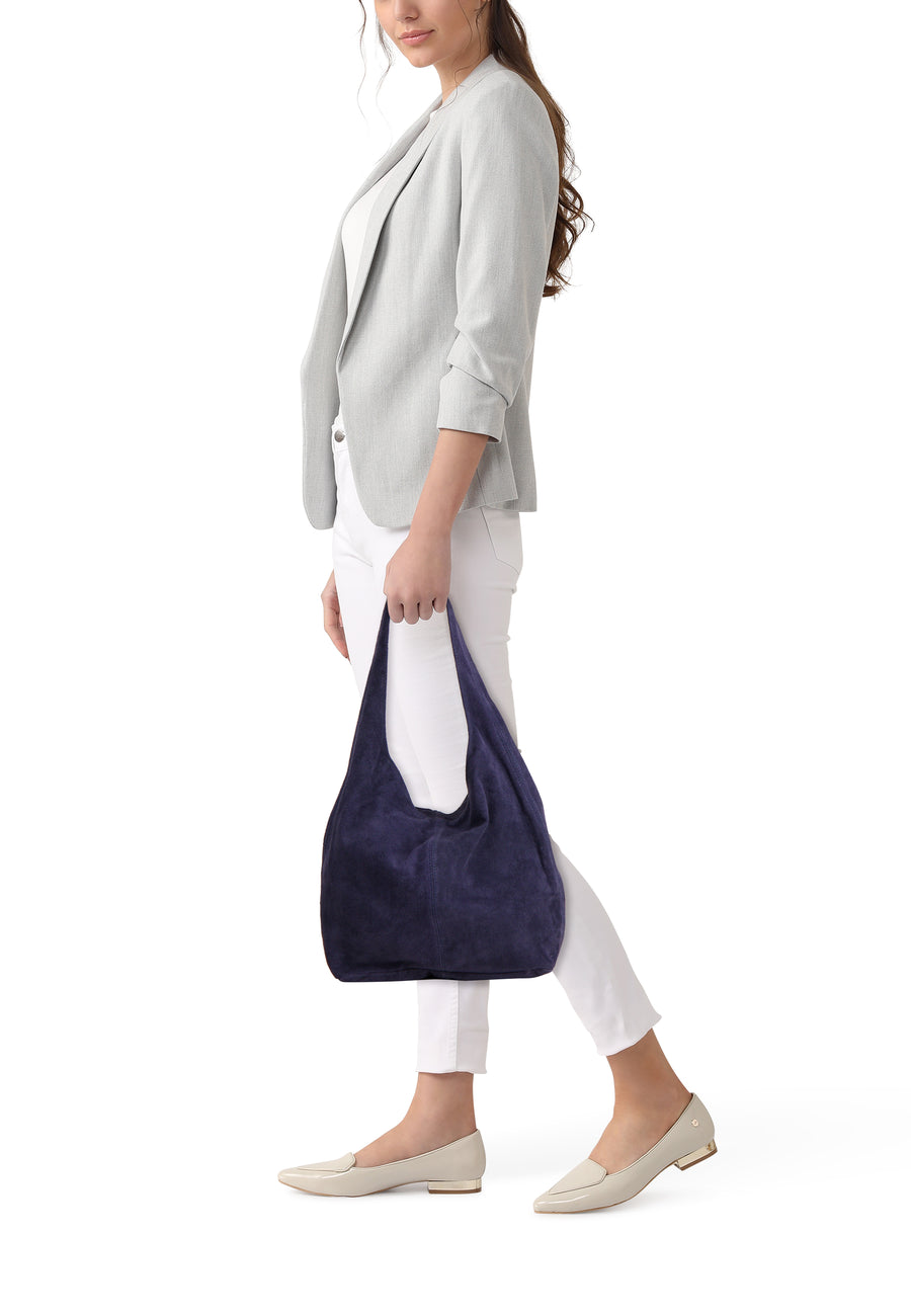 Navy blue suede shoulder bag, side view, carried by model in white pants and light gray blazer, beige loafers visible.